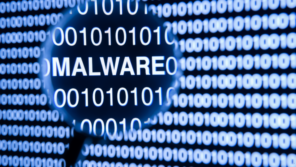 The word malware on a monitor emphasized in a magnifying glass surrounded by code.